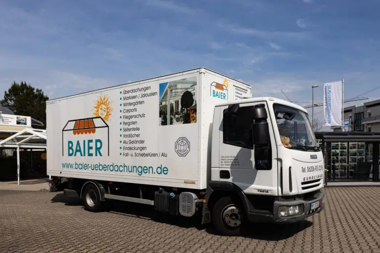 Weißer Lkw mit "BAIER"-Logo, Dienstleistungen, Banner und Gebäude im Hintergrund auf Parkplatz.