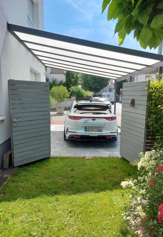 Ein weißes Auto parkt unter einem modernen Carport neben einem blühenden Rosenstrauch.
